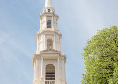 The white steeple of Park Street Church on a sunny day against a blue sky. There is a light green tree on the right side of the steeple.