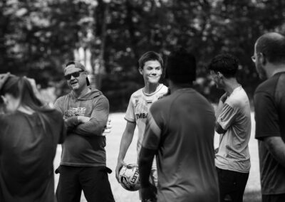 A black and white photo of an intergenerational group outdoors on a soccer field. There is a teenage boy smiling in the center holding a soccer ball. A man in sunglasses and a backwards cap stands next to him, arms crossed, talking to someone.