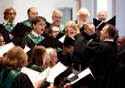 A choir wearing black robes with green stoles responds enthusiastically to a gesticulating choir director.
