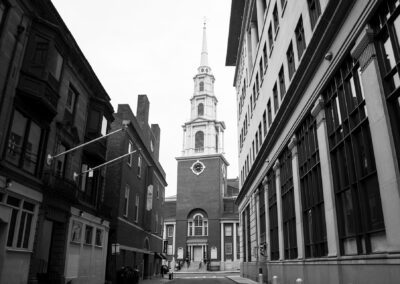 Black and white photograph of Park Street Church in Boston, shot from street level looking up at the brick church building and tall white steeple, framed by surrounding historic buildings on either side.