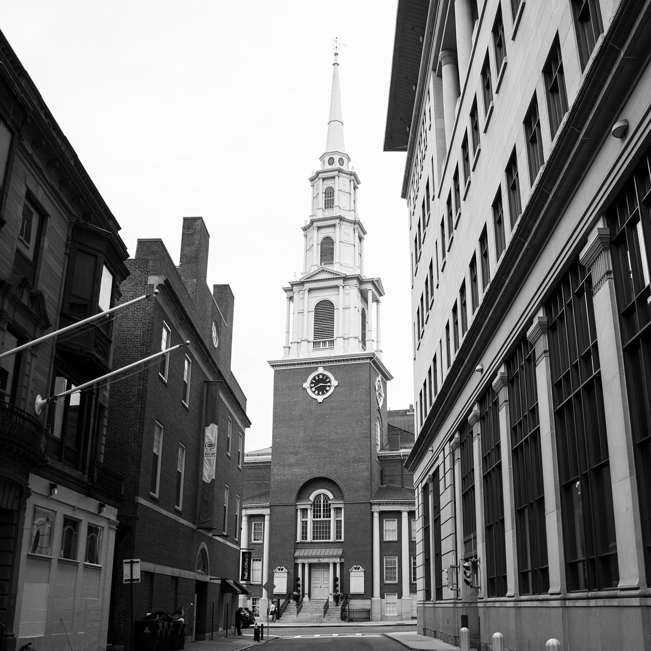 Black and white photograph of Park Street Church in Boston, shot from street level looking up at the brick church building and tall white steeple, framed by surrounding historic buildings on either side.